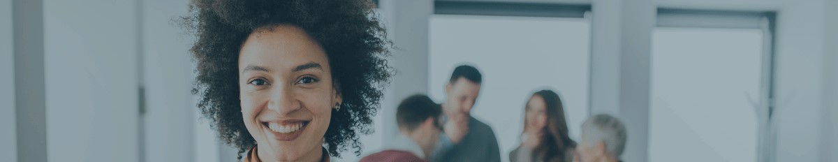 A woman looking at the camera and smiling in an office with her team members behind her talking business