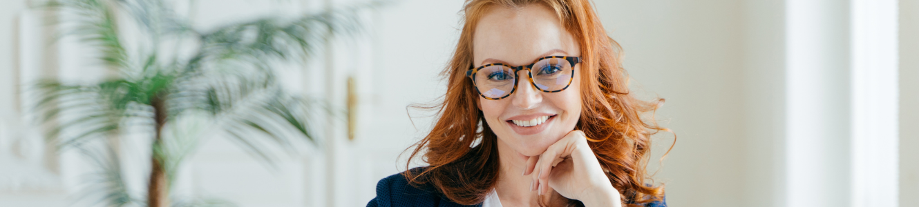 Women with red hair and glasses sitting at a desk and smiling for the camera with her hand on her chin