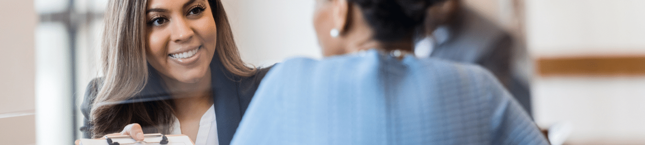 Two women talking at a table and one of the women is showing a clipboard with a paper on it