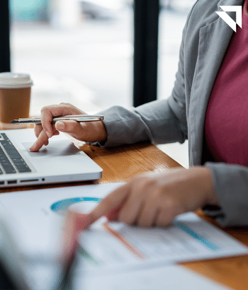 Women on her laptop scrolling and comparing as she is point at a graph that she has next to her 